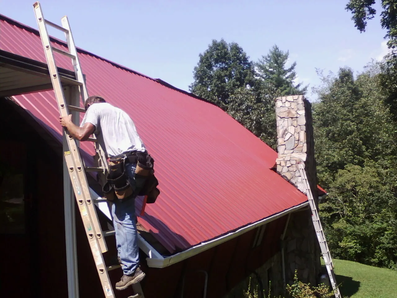 Roofer on a ladder beside a red metal roof and stone chimney — Soffit & Fascia Repair by Roofing Brothers in Cleves